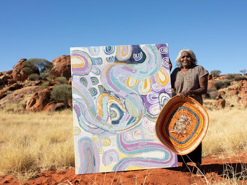 Puna Yanima with one of her baskets for Tjanpi Desert Weavers and her painting Antara (2022). Photo by Tim Coad, courtesy the artist and Mimili Maku Arts, DAAF 2022